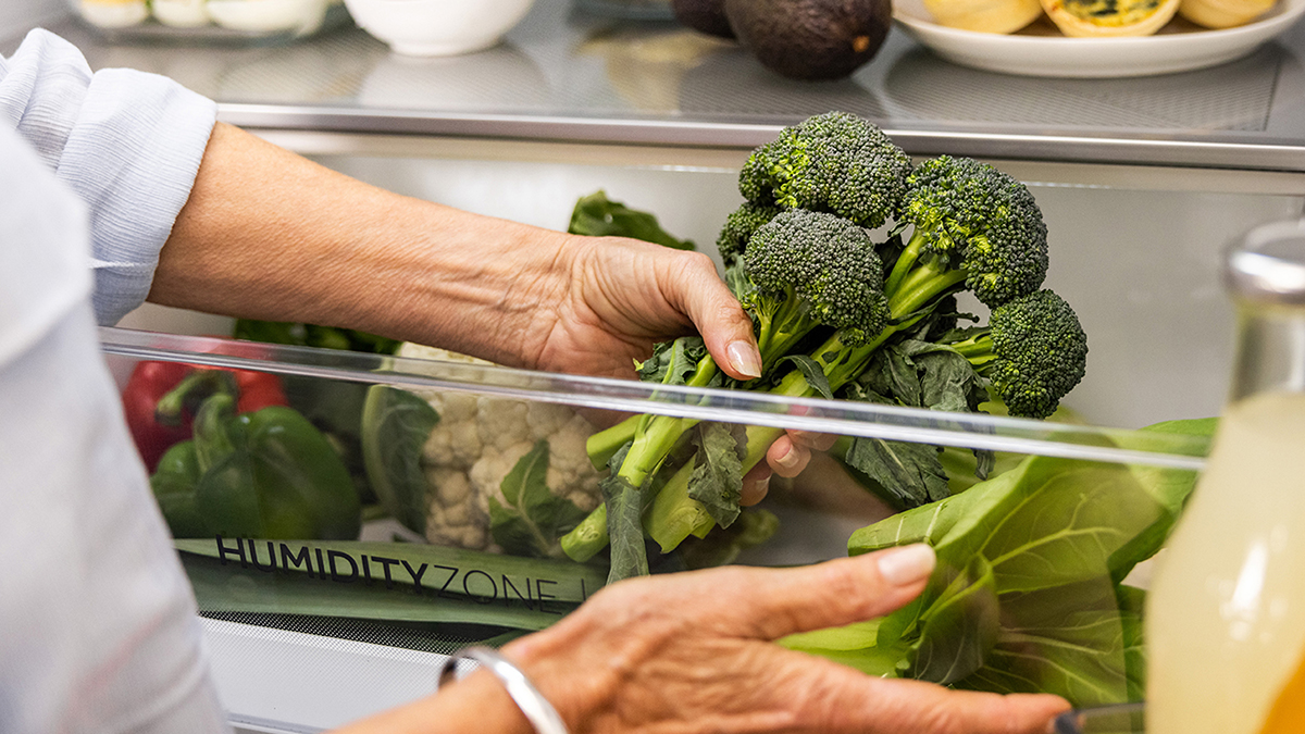 Inside a refrigerators vegetable drawer showing a hand holding broccoli
